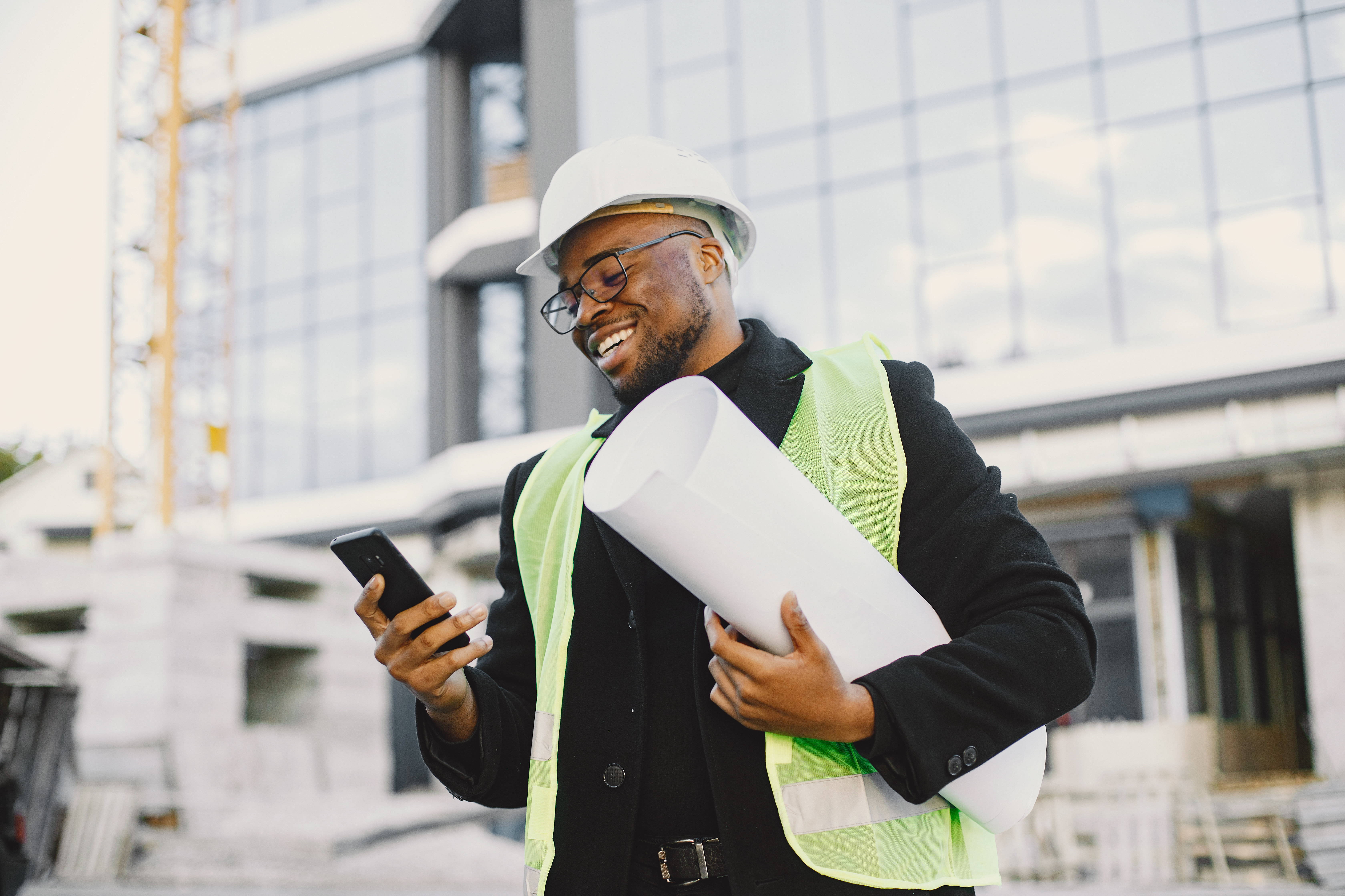 Construction professional in a hard hat and safety vest reviewing a blueprint at a building site under construction.